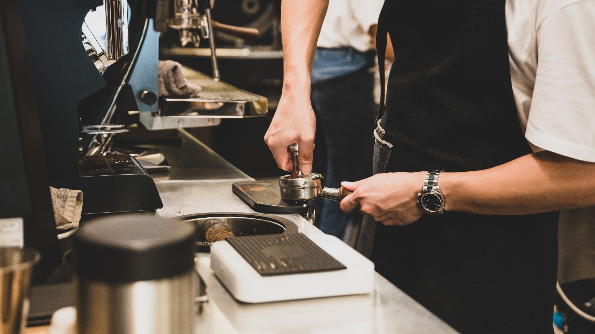 barista wearing white shirt and black apron grinding coffee on white counter