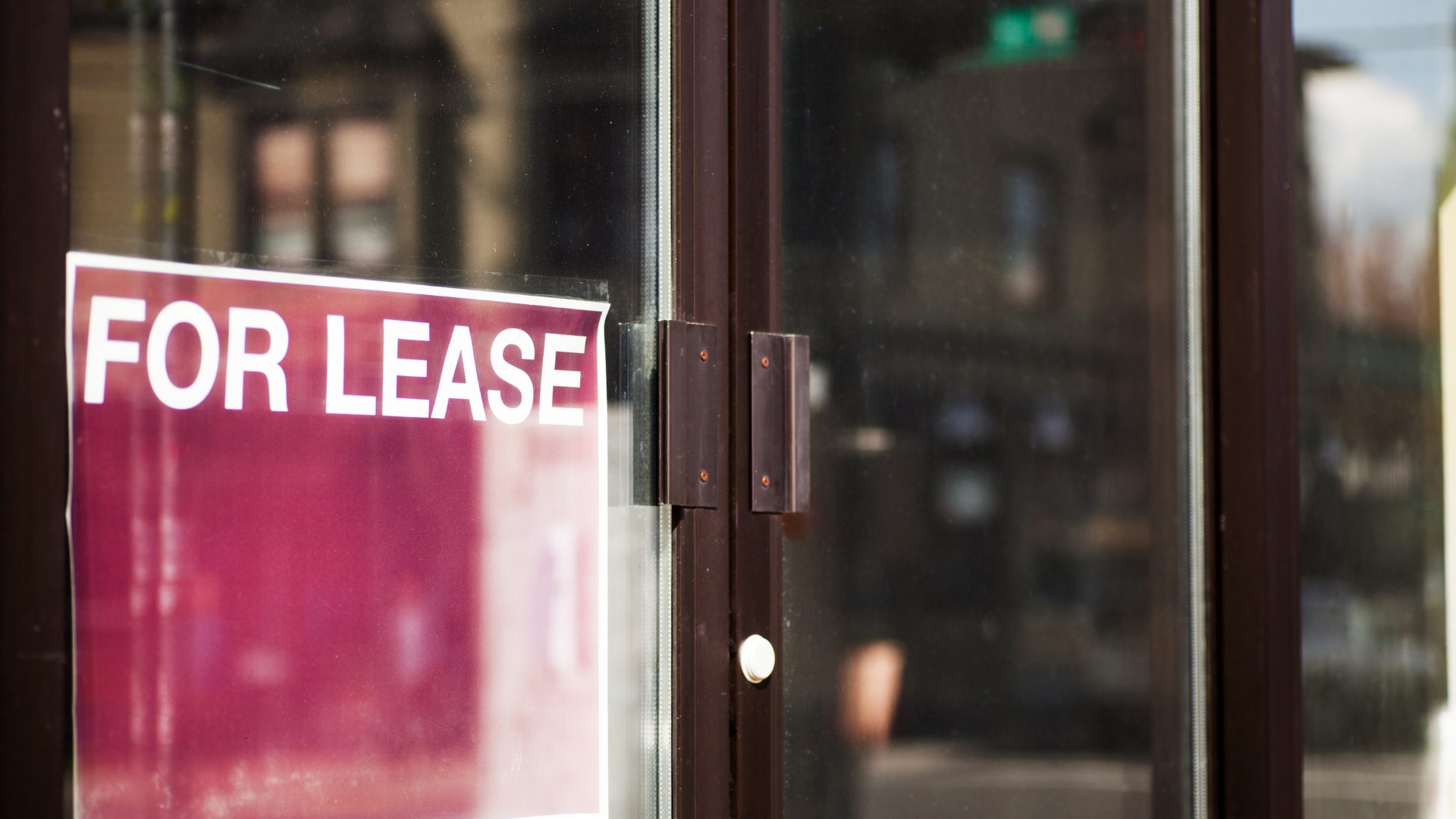 A double-door entrance to a commercial space, with a burgundy-red paper sign saying "FOR LEASE" in bold white text in all-caps