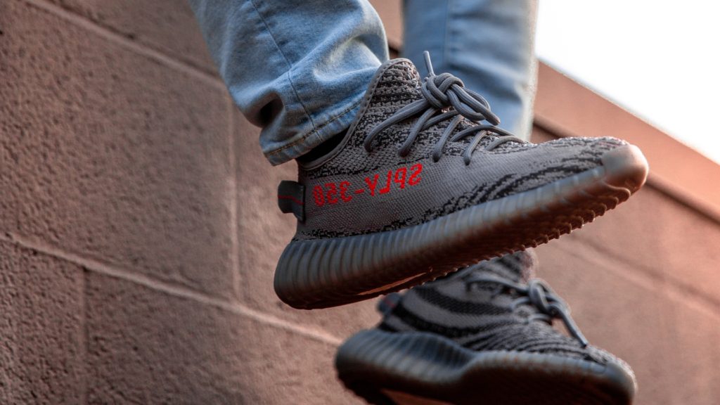 two feet dangling off of a light brown brick wall, wearing light blue jeans and gray Yeezy sneakers with bright red lettering