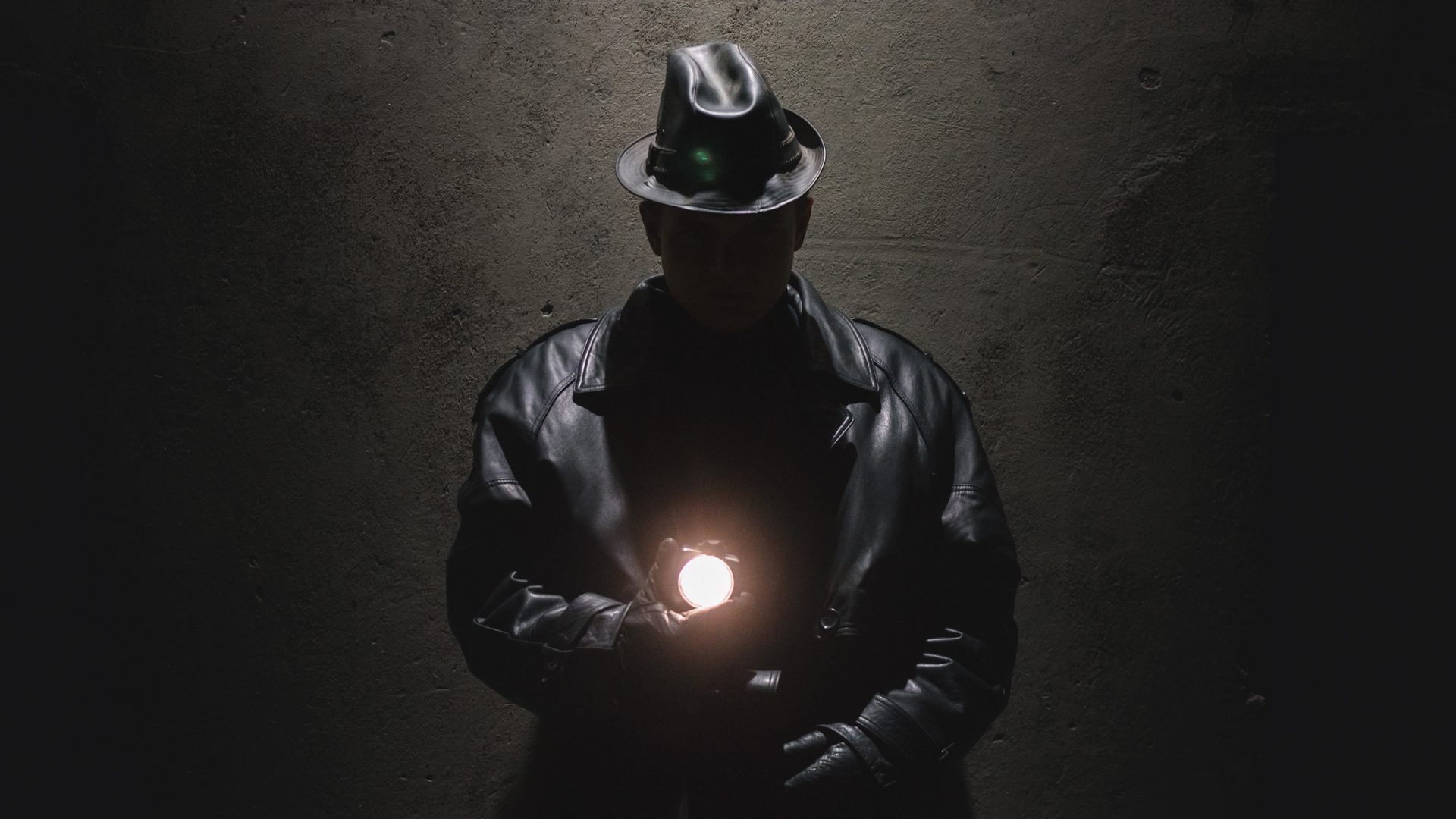 Man standing in shadows against a concrete wall. He is holding a warm-toned light in one hand in front of his chest and wearing a black leather jacket with a matching hat. He is staring into the camera and his face is barely visible