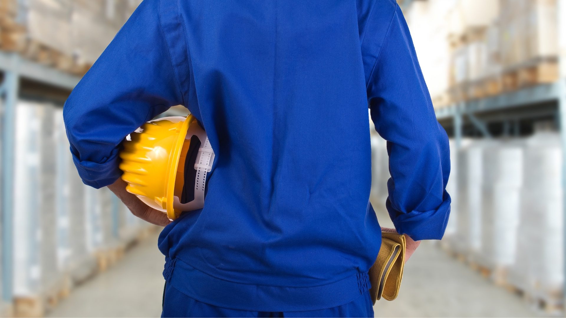 worker wearing a blue jumpsuit facing away from the camera and tucking a yellow hard hat under their left arm