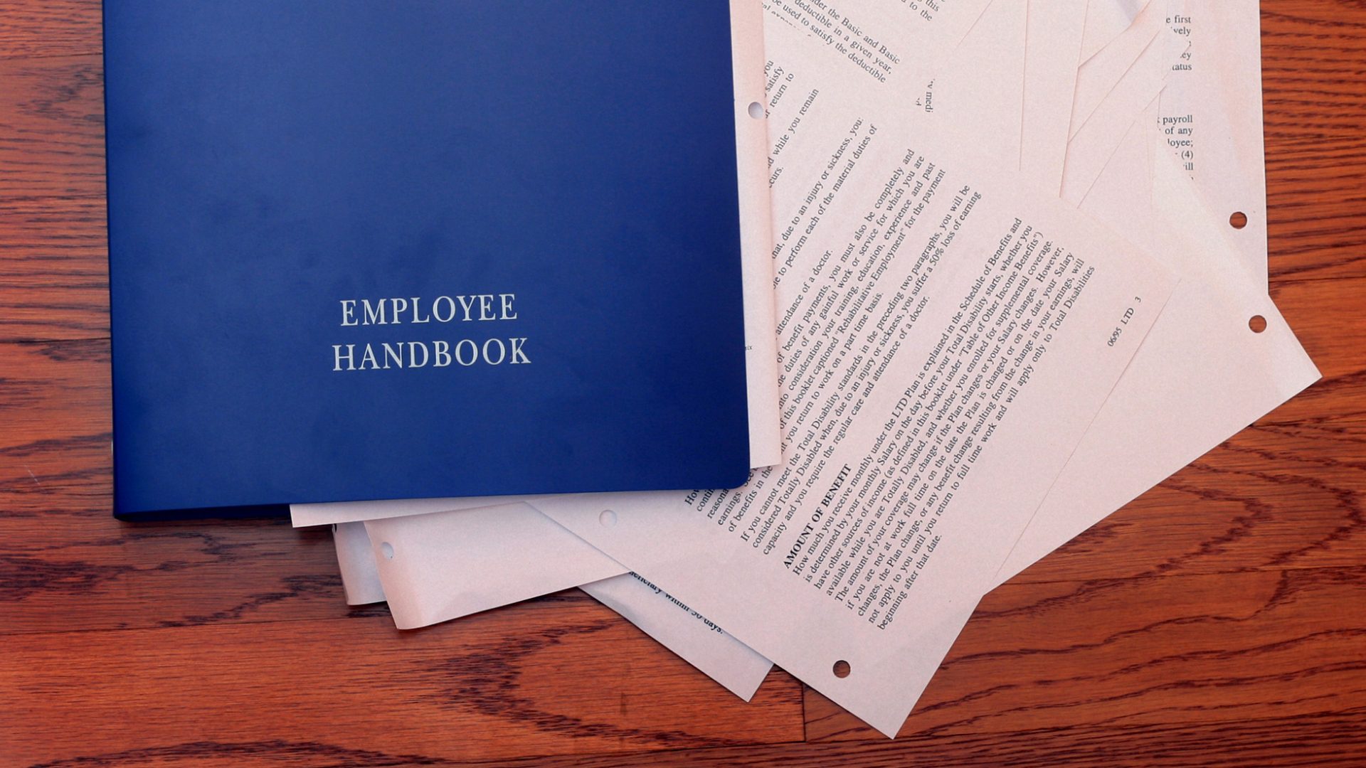 A blue book with "EMPLOYEE HANDBOOK" on the front cover lies on top of a messy stack of papers on a wood surface