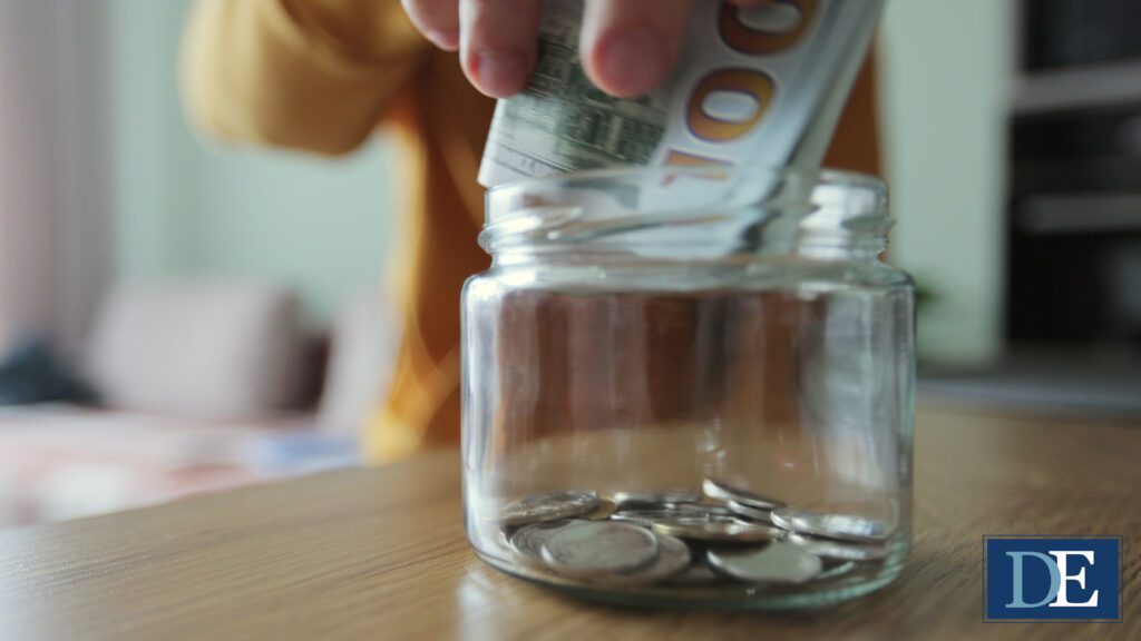 A person wearing a yellow top, unfocused in the background of the image, puts a 100-dollar bill into a clear glass jar sitting on a finished wood table. There are already several coins in it. Emblem in bottom right corner of image reads "DE" in a dark blue rectangle with a light blue D and a white E