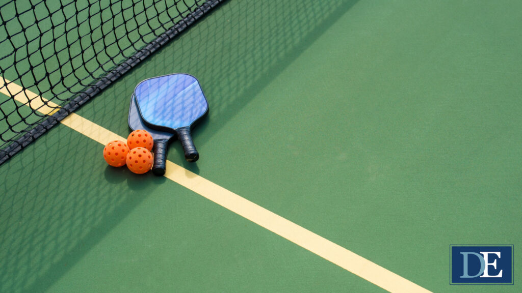 Two blue pickleball paddles and three orange pickleballs sit on the white line bisecting a green pickleball court, right next to the black net in the middle. The net is casting a shadow over the paddles and balls. Emblem in bottom right corner of image reads "DE" in a dark blue rectangle with a light blue D and a white E