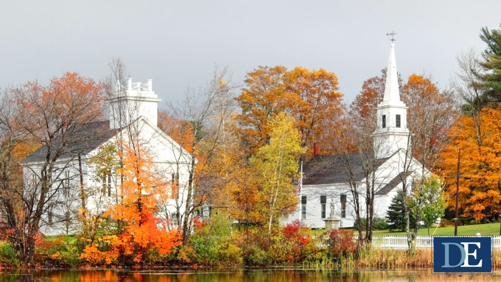 Two white colonial-style buildings sit on a grass field next to a pond with fall foliage, separated from the pond by a white fence. The sky is a light shade of gray and the shorter building is partially obscured by a tree with bright orange leaves. Emblem in bottom right corner of image reads "DE" in a dark blue rectangle with a light blue D and a white E
